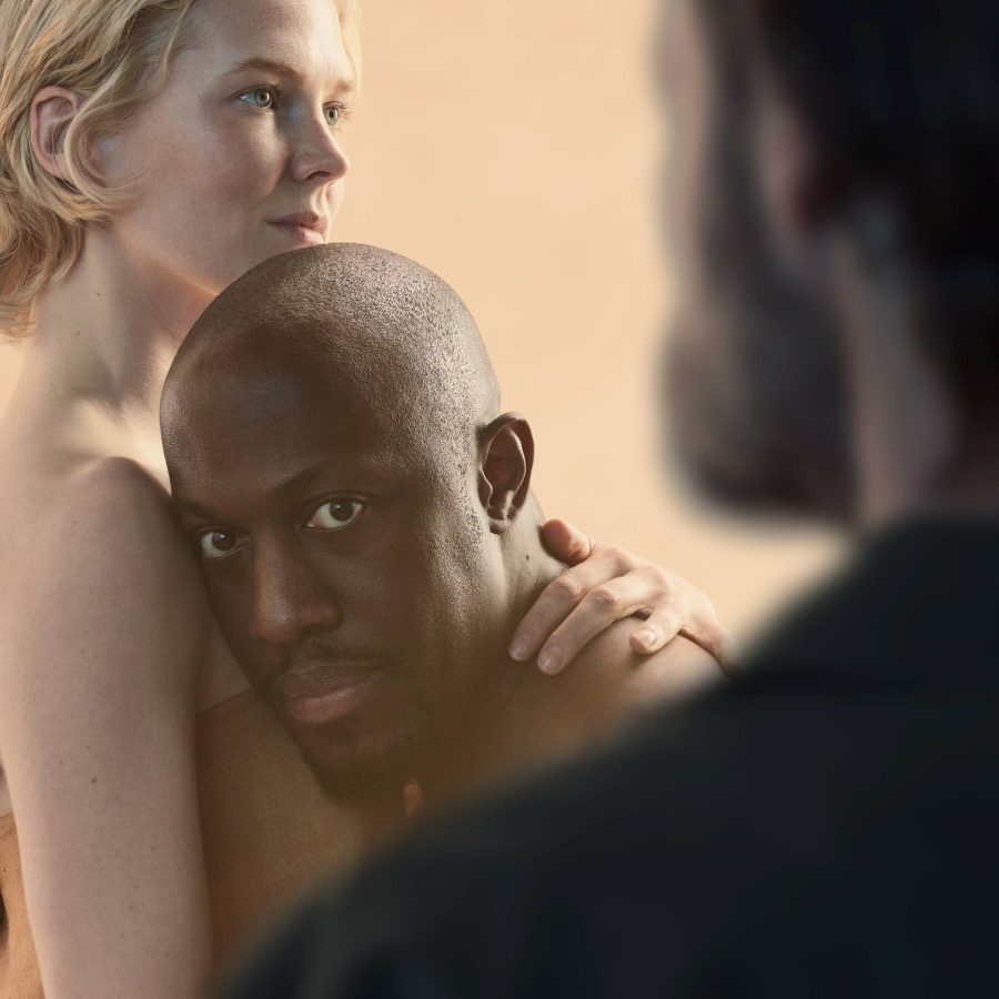 Othello, with Rosy McEwen and Giles Terera holding each other, with Paul Hilton out of focus in the foreground