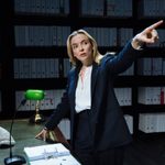 A woman (Jodie Comer) stands at a desk in a room lined with box files on shelves. One hand rests on the desk and the other is raised and pointing to something.