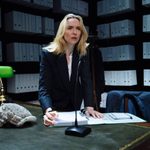 A woman (Jodie Comer) stands at a desk in a room lined with box files on shelves. She holds onto a book and leans towards a microphone on the desk.