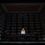 A woman (Jodie Comer) sits on a desk in a room lined with box files on shelves.