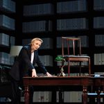 A woman (Jodie Comer) stands at a desk in a room lined with box files on shelves. She leans on the table which upon it sits a chair.