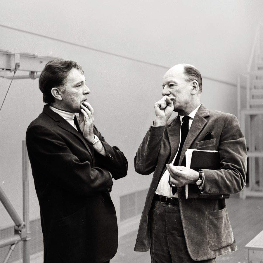 A black and white photograph of Richard Burton and John Gielgud, standing face-to-face, in mid-conversation, each holding a hand to their chin.