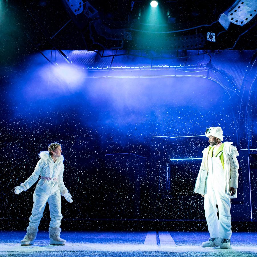 Two people in white, winter attire stand on a snow-covered stage beneath blue and purple lights. They face each other, surrounded by falling snow, creating a dramatic and immersive scene.