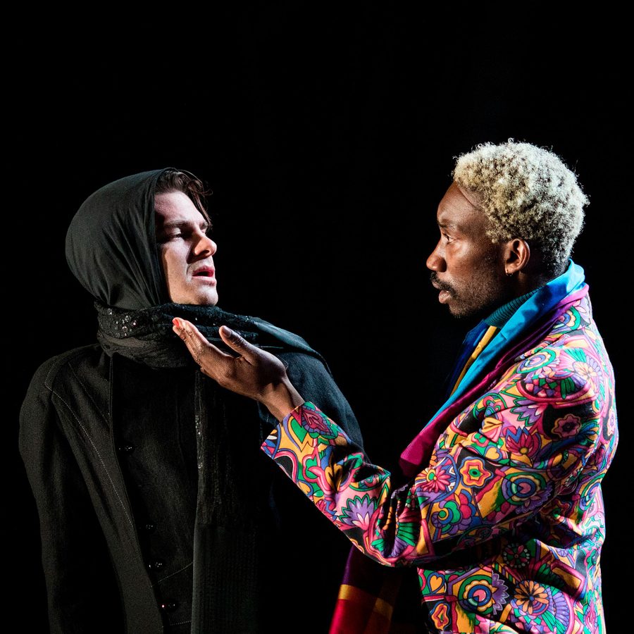 Two actors in a powerful scene: one is dressed in black with a hood, looking serious, while the other, with blond hair, wears a vibrant, colorful jacket, gesturing expressively against a dark background.