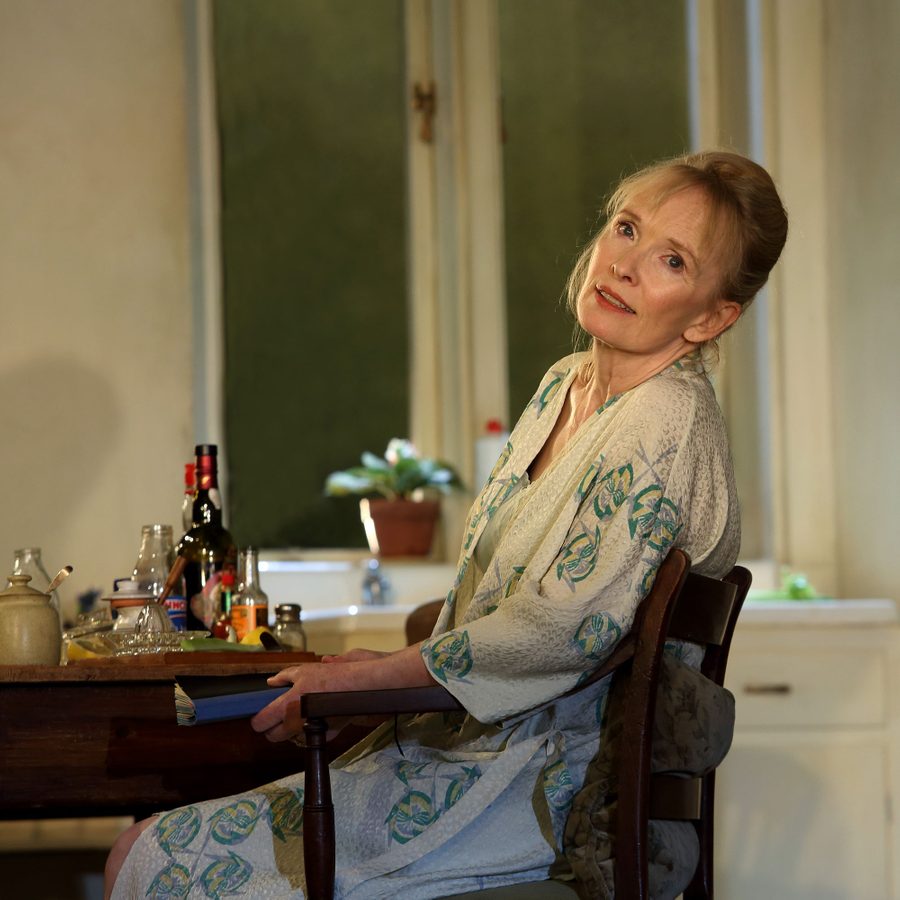 A woman sitting at a kitchen table, leaning back slightly. The table has various bottles and a pot. She wears a light-colored, patterned robe. The background shows tall windows with frosted glass and a plant on the windowsill.