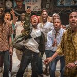 A production image from 'Barber Shop Chronicles' at the Dorfman Theatre, 2018, featuring a group of Black men all watching something with excited and shocked faces