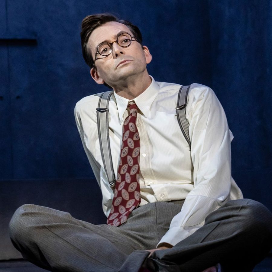 David Tennant sitting on the floor, wearing glasses, shirt, tie, braces and trousers.