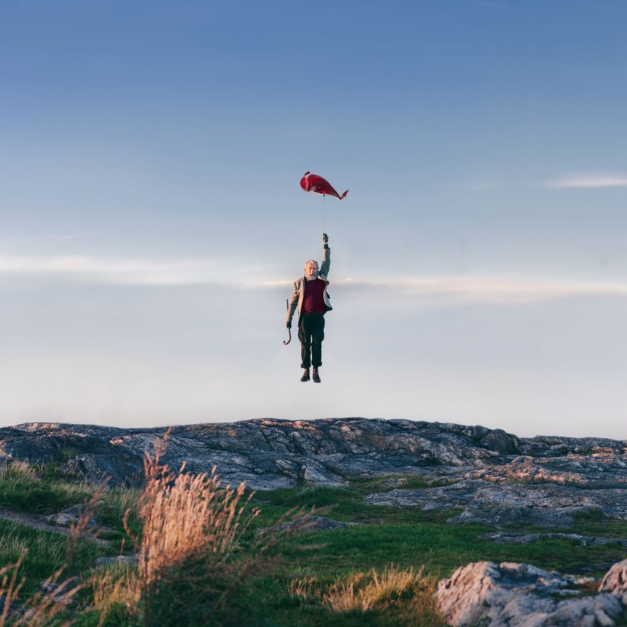 Under-Milk-Wood-Mast-Head-2000x1000 An elderly man in a suit floats above rocky terrain, holding onto a red balloon shaped like a fish against a blue sky with scattered clouds.