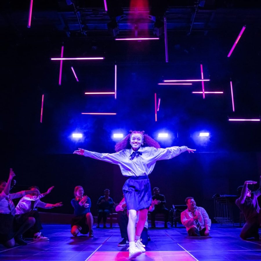 Alex Jarrett steps forward, centre-stage front, arms outstretched and smiling, with members of the company sat or crouching behind her.