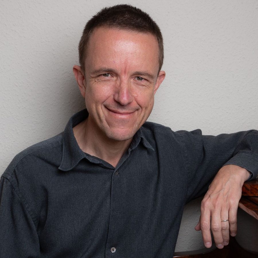 A man sits, leaning against a wall, one arm on a piano-lid, smiling straight to camera.