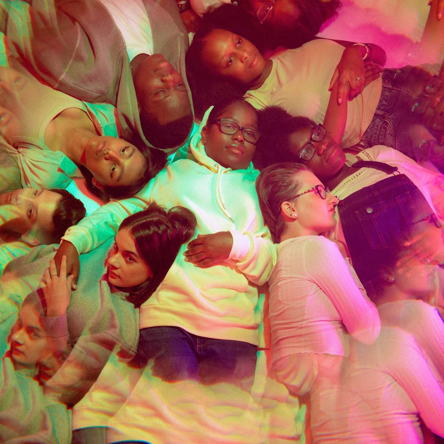 A group of young people rest in a circle, looking out and away from the group, with a soft pink and green glow effect over the image. One young person in the centre wears glasses and looks directly into the camera. The National Theatre logo and Connections 2023 logo are written at the bottom of the image in white.