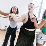 Three women in athletic wear practice a dance routine in a brightly lit studio, pointing forward in unison. Behind them, large windows and wall-mounted ballet barres are visible. Three fans are placed on the floor in the background.