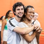 Two people are smiling and embracing in a dance studio with a bright orange wall. They stand close to one another, with the person behind holding the hands of the person in front. Other people in various dance poses are visible in the blurred background.