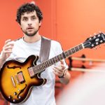 A man with curly hair and a beard is playing an electric guitar. He is wearing a white shirt and a guitar strap over his shoulder. The background is a bright orange wall with some furniture partially visible. The man appears to be speaking or explaining something.