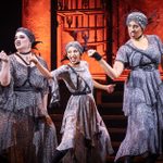 Three performers on stage wear coordinated, patterned costumes and headbands, striking animated poses under warm lighting. A dramatic set featuring a tall, ornate backdrop is visible, enhancing the theatrical atmosphere.