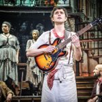 A performer stands center stage holding a guitar, wearing a white apron and red neckerchief. Three other performers watch him from different positions on stage, dressed in rustic costumes. The background features a dimly lit, multi-level set.