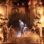 A dramatic stage scene with performers in rugged costumes, illuminated by warm, smoky lighting. The central character stands on a raised platform, surrounded by others. The background features an ornate balcony and atmospheric fog.