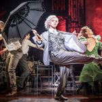 A performer in a gray suit and white hair dances energetically on stage holding a black lace umbrella. Another dancer in a green dress joins in. The vibrant ensemble creates a lively scene with varied costumes and dramatic lighting.