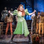 A woman in a vibrant green dress energetically sings on a dimly lit stage, holding a bottle in each hand. She is surrounded by actors in casual attire and musicians, with a dramatic backdrop of stairs and instruments.