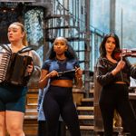 Three women stand in a rehearsal space. One plays an accordion, another holds a small object, and the third plays a violin. They are surrounded by musicians and stage equipment. The room has a rustic and artistic ambiance.