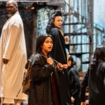 A group of actors in costume are on a stage set. A young woman in the foreground looks upward with a curious expression, wearing a dark coat and backpack. A man in a white robe and a woman in a black coat stand behind her. Stairs and set pieces are visible.