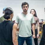 A group of people stand closely together in a circle, appearing serious. The person in the center, wearing a light gray shirt and jeans, looks forward with determination. Others, dressed casually, face inward, focused on the central figure.