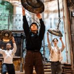 Three people hold large metal lampshades hanging above them, in a rustic, industrial setting. The room features a balcony with ornate railings, wooden floors, and exposed pipes. They appear focused, rehearsing for a performance.