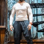 Man in a white long-sleeve shirt and dark tracksuit bottoms stands confidently on a stage with a spiral staircase in the background. He wears a red watch and light-colored sneakers. The setting has a vintage, industrial feel.