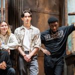 Four people stand casually on a set that resembles a rustic room, with wooden and metallic elements. One person sits while others lean against objects. They are dressed in casual attire, and the setting has an industrial feel.