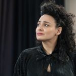 Person with curly black hair looks to the side with a thoughtful expression. They wear a pleated black blouse, and the background is blurry with dark curtains.