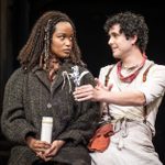 A person with long curly hair, wearing a dark textured coat, holds a white candle while looking intently at another person with short curly hair, who is gesturing and dressed in a white shirt with braces and a red scarf. They are on a dimly lit stage.