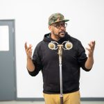 A man wearing a camouflage cap and black hoodie is standing in front of a microphone. He gestures with both hands, and is in a room with white walls and a pair of closed doors in the background.