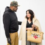 A man in a camouflage cap and dark hoodie stands next to a woman in a white fur coat holding a woven bag with a floral design. They appear to be having a serious conversation in a room with a gray door and light walls.