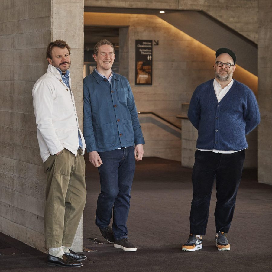 Three men stand together and smile at the camera. One wears a white shirt, one a blue shirt and the other a navy cardigan. The backdrop is concrete walls and the top of a staircase.