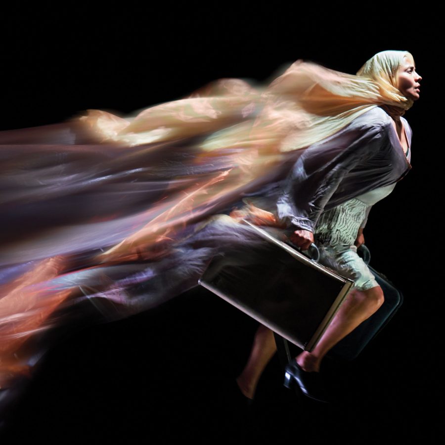 A woman runs, carrying a black suitcase with metal trim, her coat and headscarf billowing and blurred, behind her.