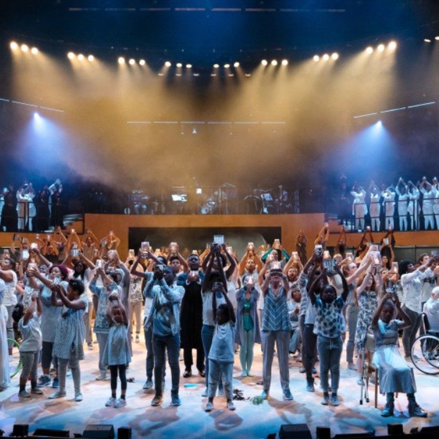 Pericles-candles-2000x1000-JamesBellorini A large community ensemble onstage all lifting candles and dressed in white and cream colours