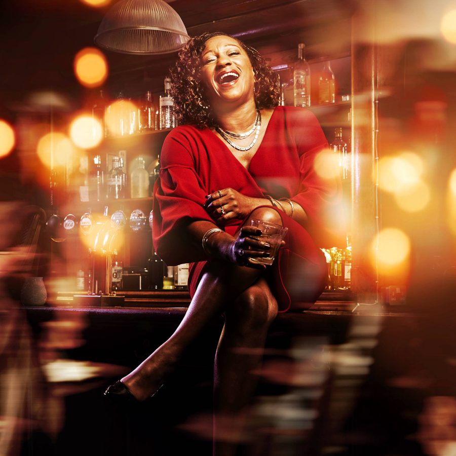 A laughing woman wearing a red top, holding a drink in a bar, surrounded by spots of light.