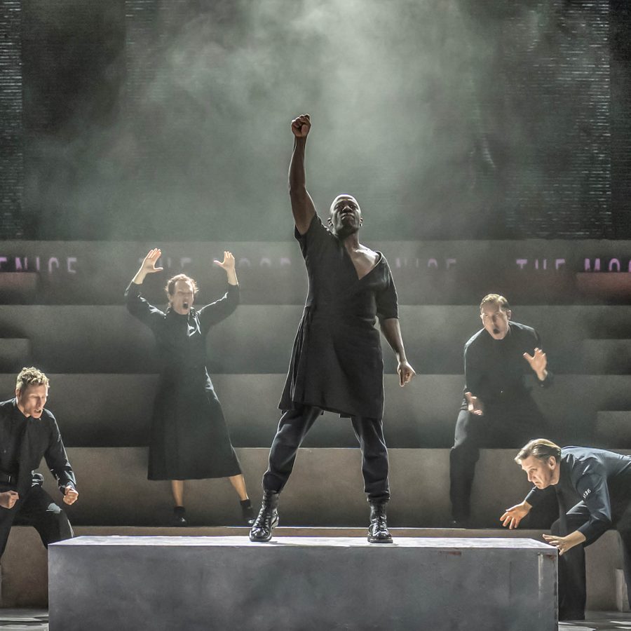 NTGDS_ES_NTatHome_Othello_TakeYourSeats_Web_2000x1000 Othello stands centre stage on a raised platform, his fist raised, surrounded by figures in expressive poses.