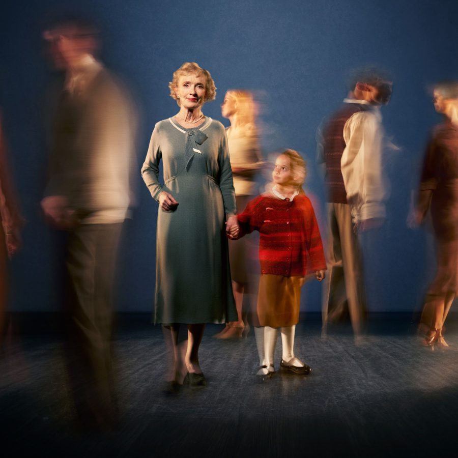 A woman in a brown long-sleeved dress stands, smiling, looking straight to camera. She is holding hands with a young girl who looks up at her. The blurred figures of five other people stand either side of them and the girl is also blurred.