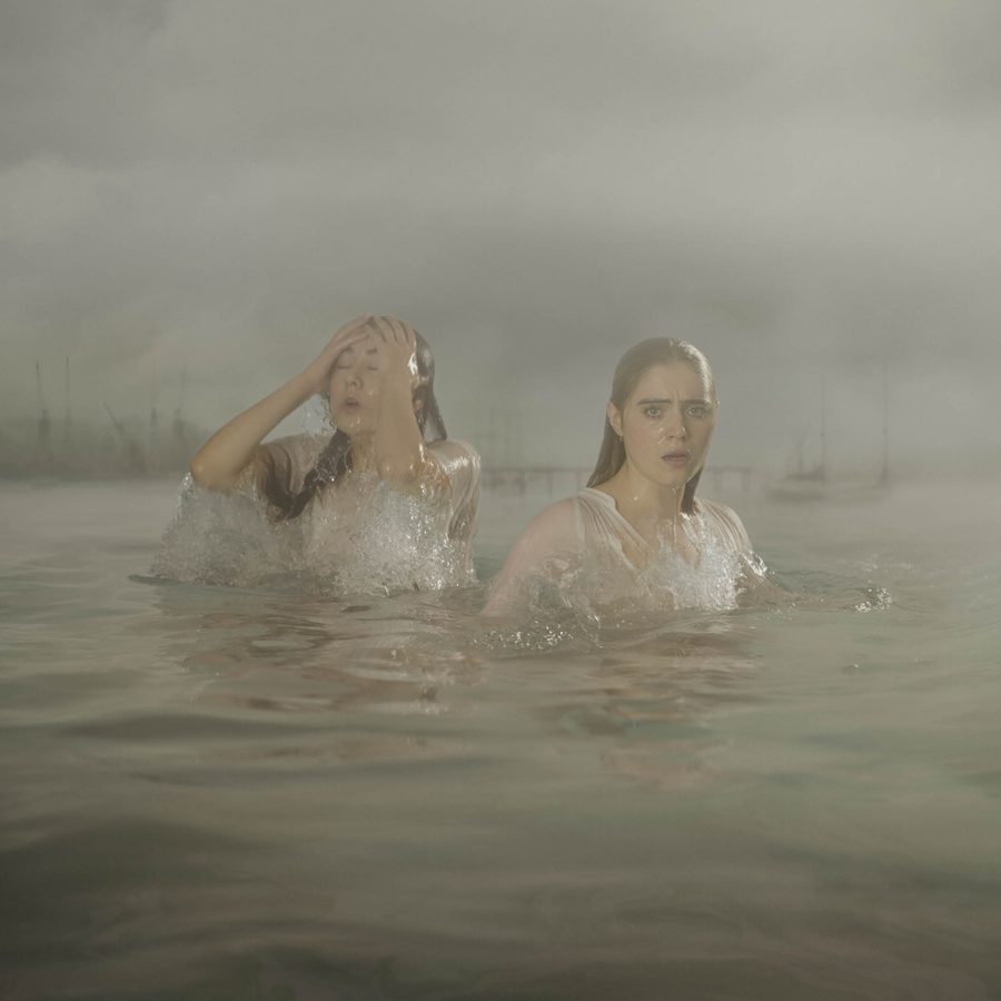 Two women in thin white dresses stand in a river, soaking wet, one is pushing her hair back from her face. A low-lying city skyline and bridge are behind them in the distance.