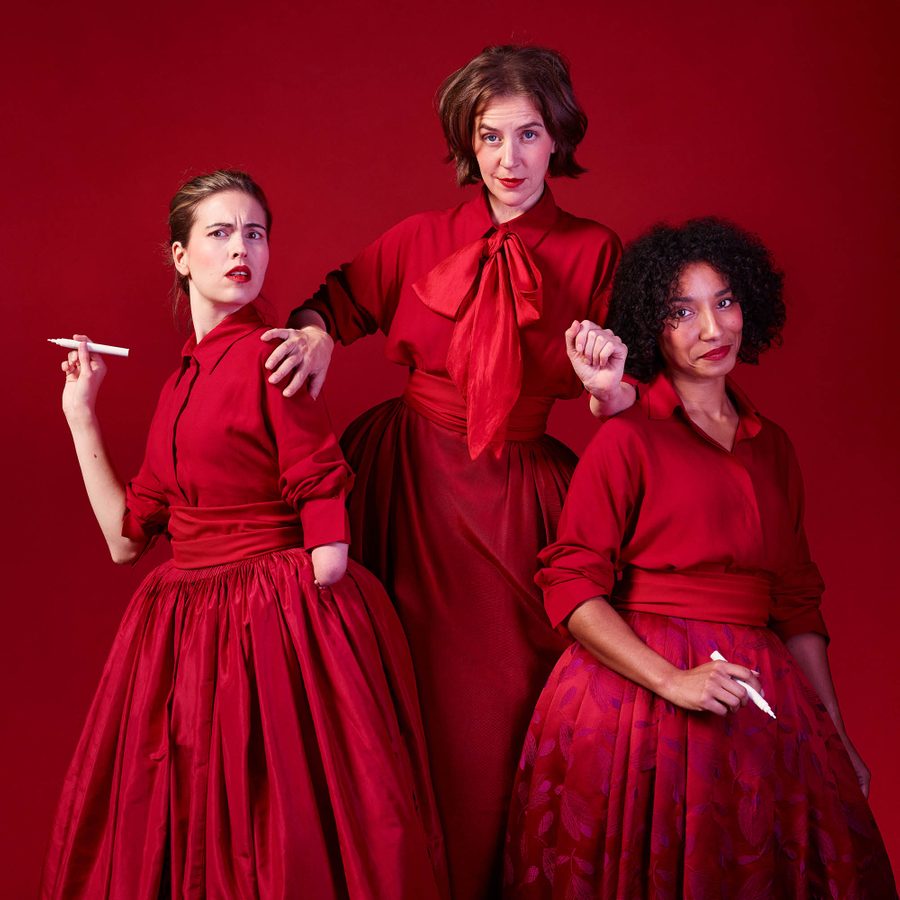 Three women stand together, all facing to camera, the middle one, slightly higher than the others, seems to be trying to push the others, who each hold a pen, aside. All wear red shirts and skirts and the middle one has a large red bow tied at her collar.