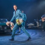 A (Oliver Chris) man wearning a patterned blue suit, strides across stage, a bunch of flowers in one hand and a teddy bear in the other. A sports car, scooter and musicians are in the background.