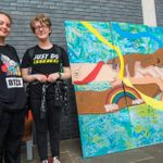 Two young people stand beside a large artwork featuring four hands embracing with peace and rainbow signs