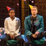 Two actors sit on a table on a stage in party hats. The person on the right wears a 