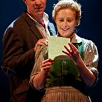 A woman in period attire happily reads a letter while a man in a hat and suit jacket stands behind her, looking over her shoulder. The background is dark, casting a spotlight on the pair, highlighting their expressions and creating an intimate atmosphere.