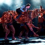 A life-sized puppet of a horse galloping and being ridden by a young man and operated by three puppet handlers.