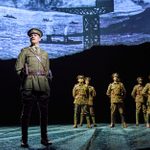 A man in a military uniform stands at attention on a stage, with five other uniformed individuals in the background. The backdrop features a monochromatic image of a shipyard or harbor. The scene is from a theatrical production.