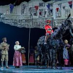 A stage production featuring several actors, with one in military uniform riding a large horse puppet. The background shows a projection of an old street scene with colorful bunting. Other actors in period clothing stand nearby, watching and interacting.