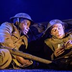 Two soldiers in World War I uniforms are sitting in a trench at night. The soldier on the left holds a rifle and looks toward the other soldier, who is reading a letter. Both wear helmets and appear to be in a serious conversation or situation.