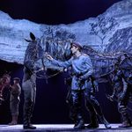 Actors on stage performing a play featuring large, lifelike horse puppets. One actor, dressed in a military uniform, interacts with a horse puppet while others dressed as soldiers look on. A backdrop displaying the words 